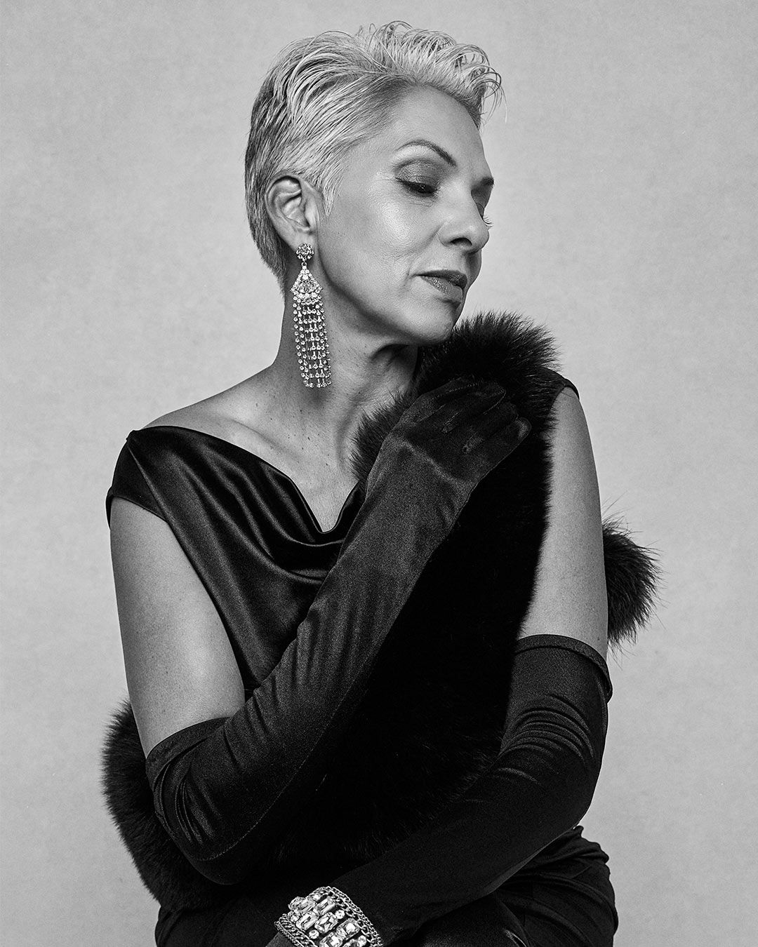 Elegant older woman in black velvet gown with fur gloves and chandelier earrings.