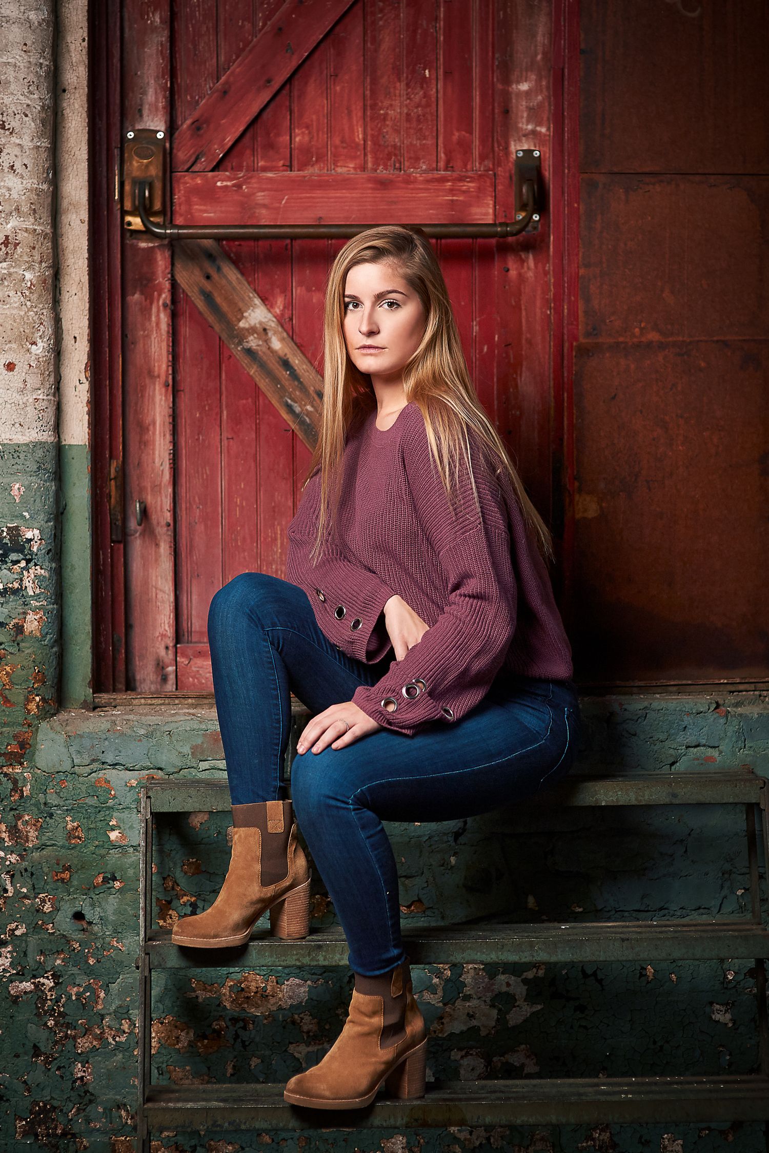 Young blonde woman in purple sweater and jeans sitting on rustic steps by red barn door.