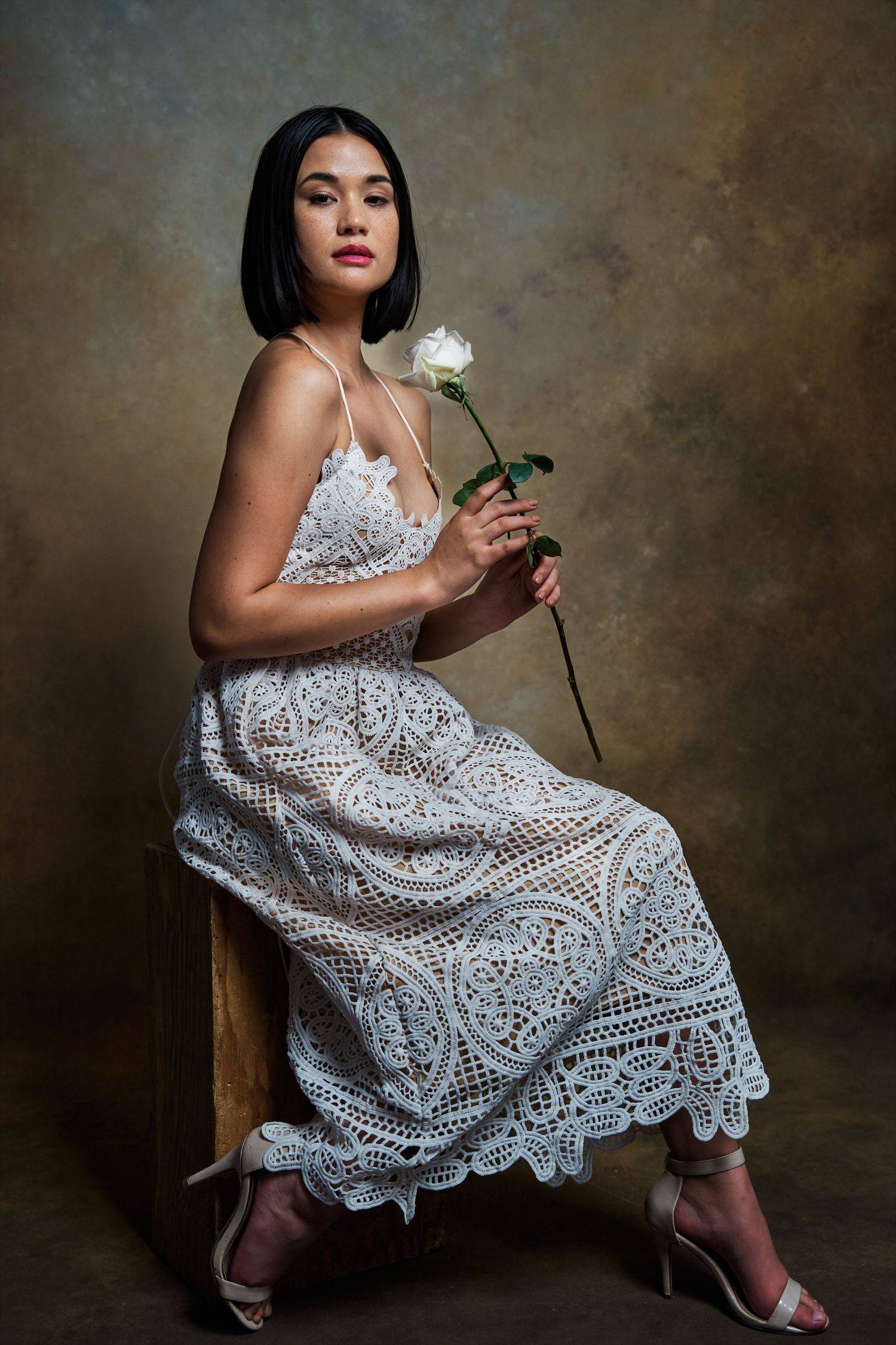 Young woman in white lace dress holding a white rose while seated.