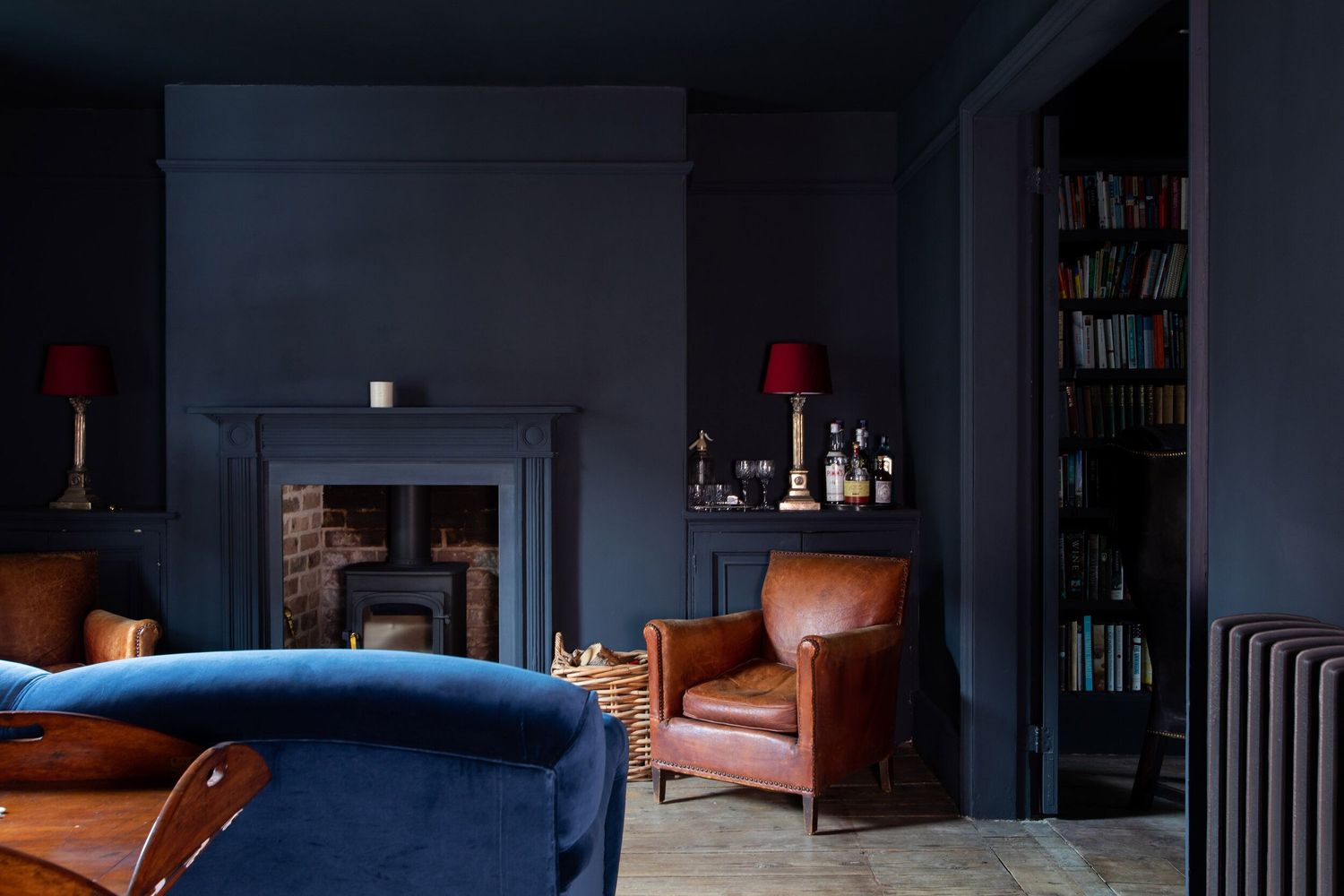 Dark blue living room with velvet sofa, leather armchair, and red lampshades creates a moody, sophisticated atmosphere.