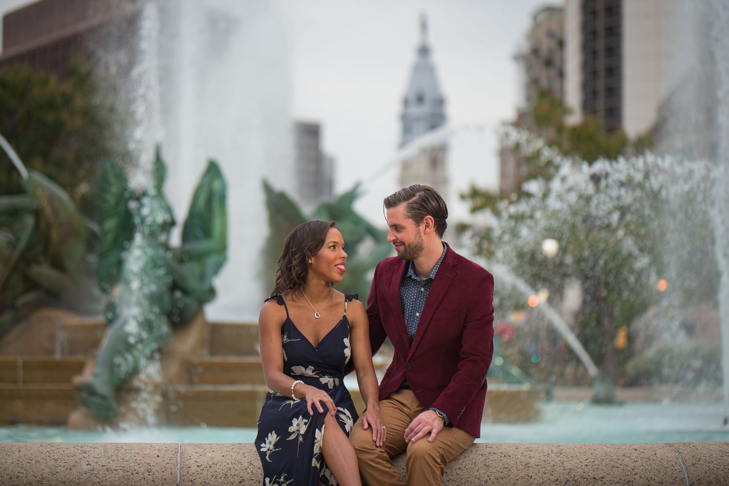 Couple enjoys romantic moment by fountain with Philadelphia cityscape in background.