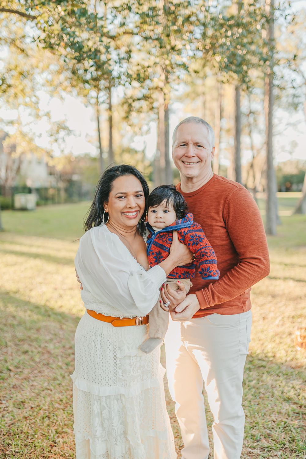 A heartwarming family portrait taken outdoors with tall trees and warm lighting.