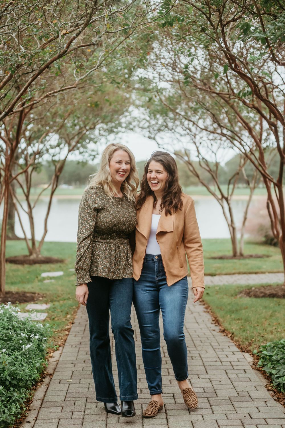 Two friends pose together on a landscaped brick pathway with trees and a lake in the background.