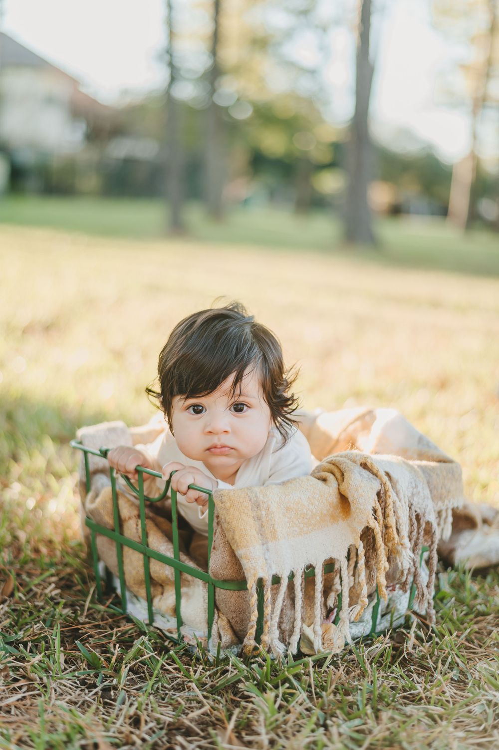 Baby in white outfit lying in rustic basket with blanket on grass.