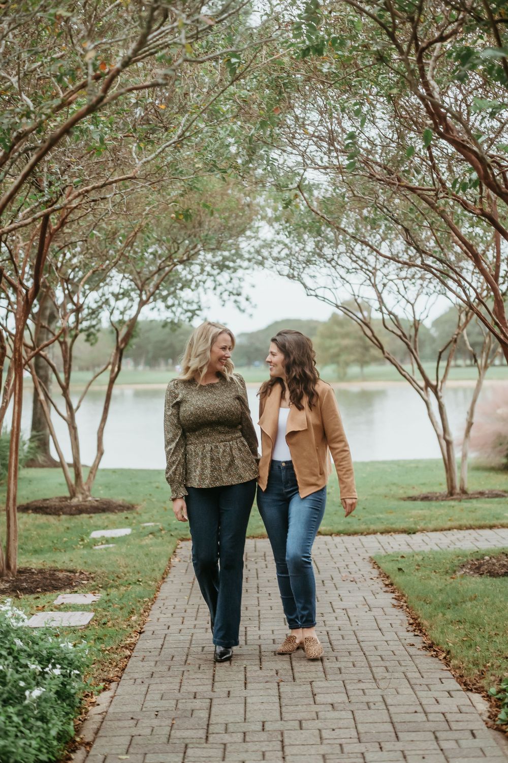 Two friends walk together on a brick pathway surrounded by trees and landscaping near a lake.