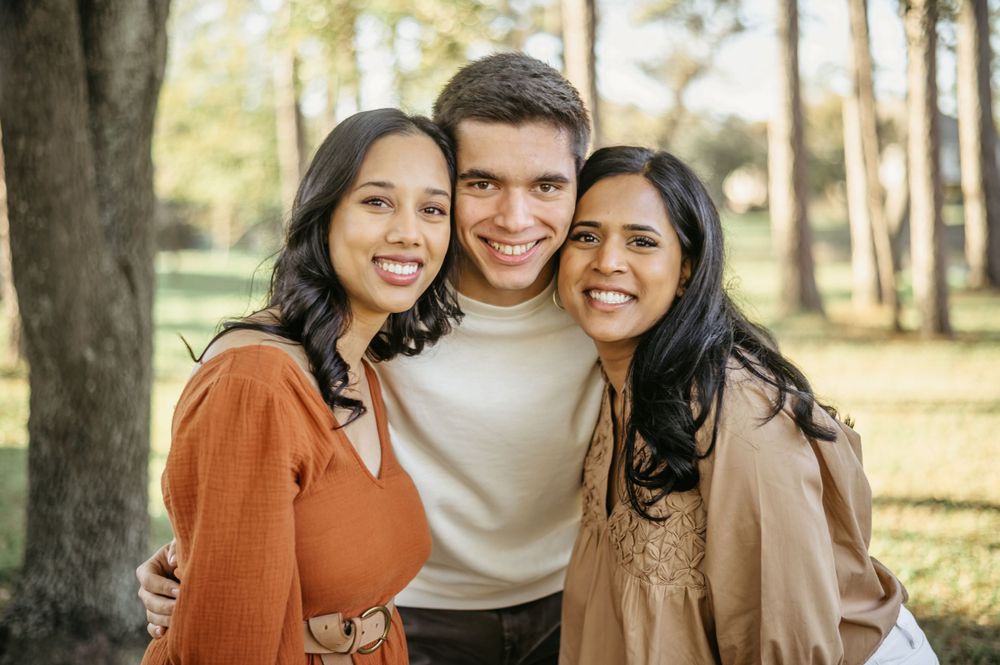 Three friends in warm-toned sweaters hugging and smiling outdoors in a park setting.