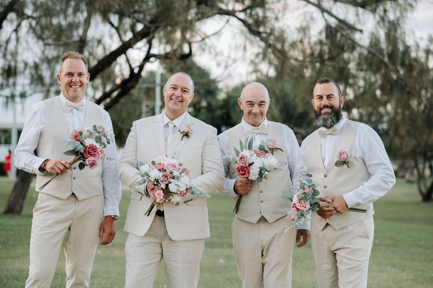 Groomsmen in beige suits hold pink floral bouquets while smiling outdoors.