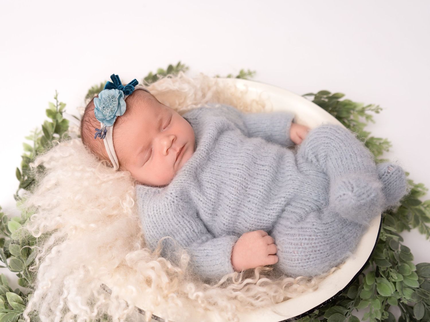 Baby girl in blue romper sleeping in a cosy fluffy bowl with foliage, at newborn photoshoot in Doncaster.