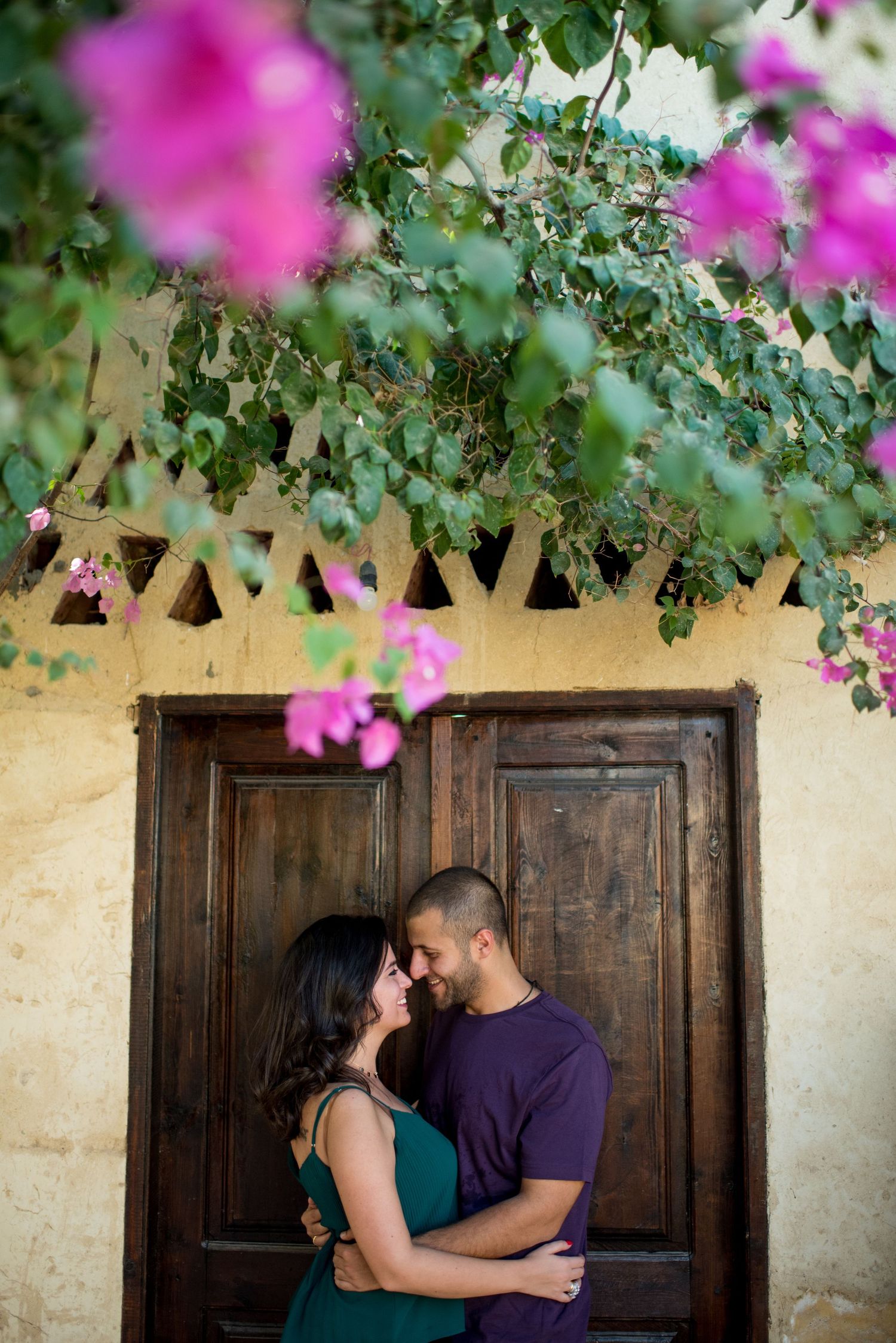 A couple embraces in front of a rustic wooden door with vibrant pink bougainvillea flowers overhead.