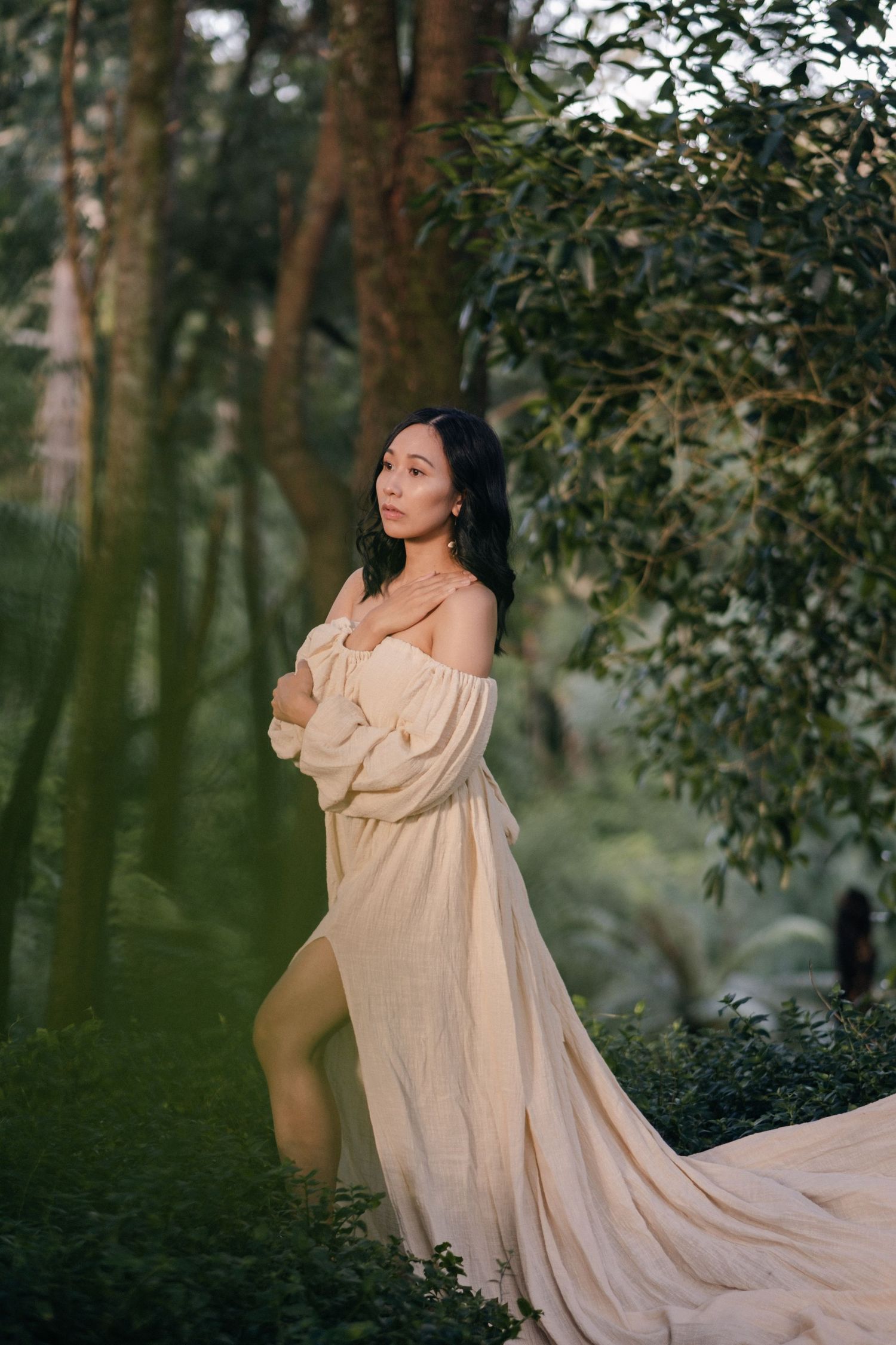 Ethereal maternity photo in flowing cream dress among forest trees.