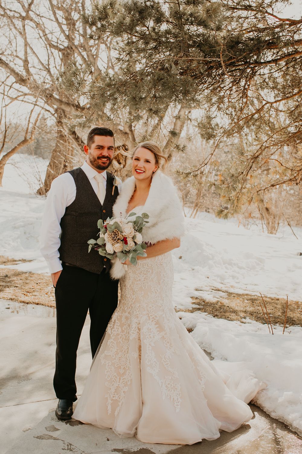 Bride in lace gown with fur wrap and groom in dark vest smile together in a snowy winter wedding setting.