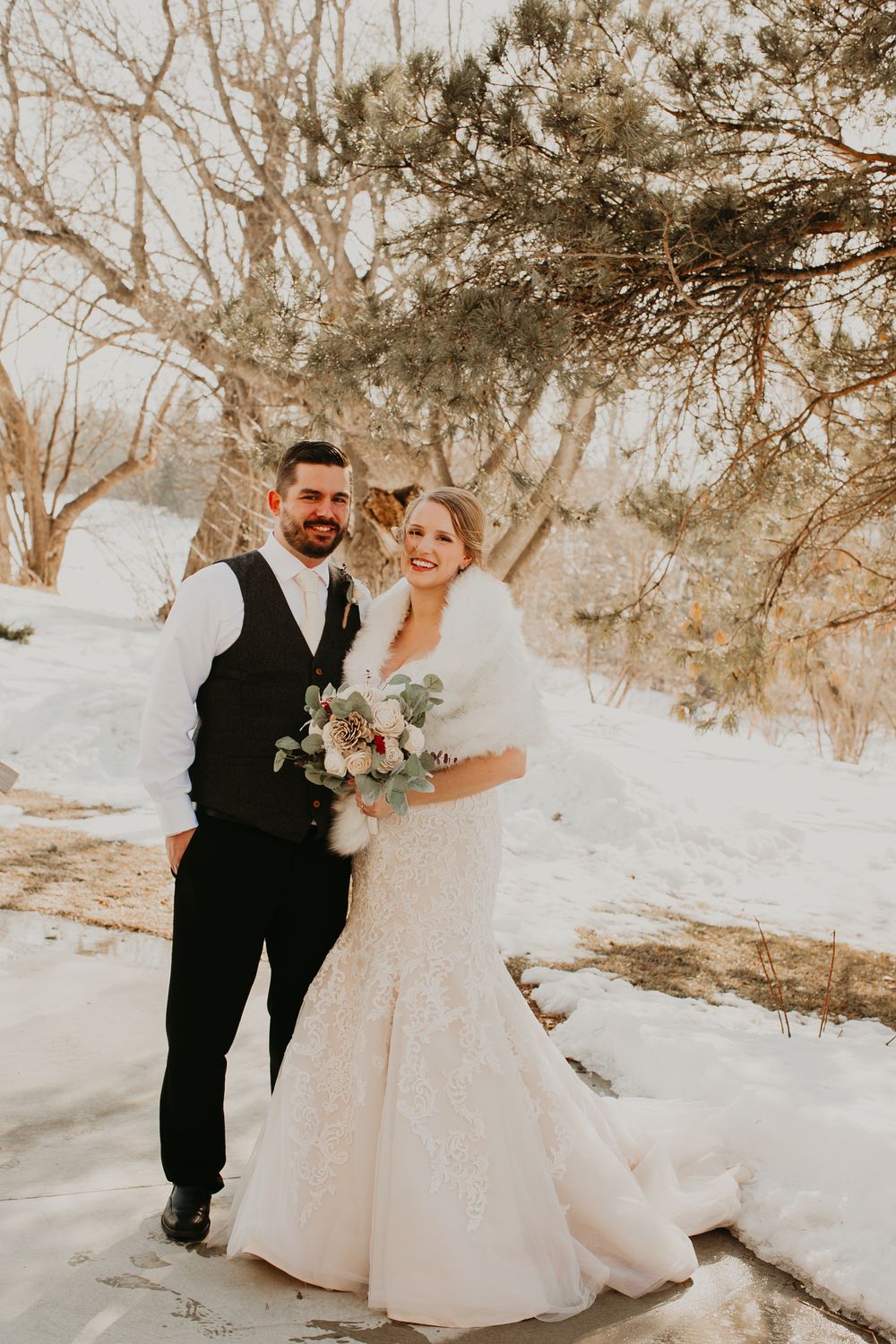 Bride and groom pose happily in snowy winter landscape, bride wearing fur wrap and holding elegant floral bouquet.