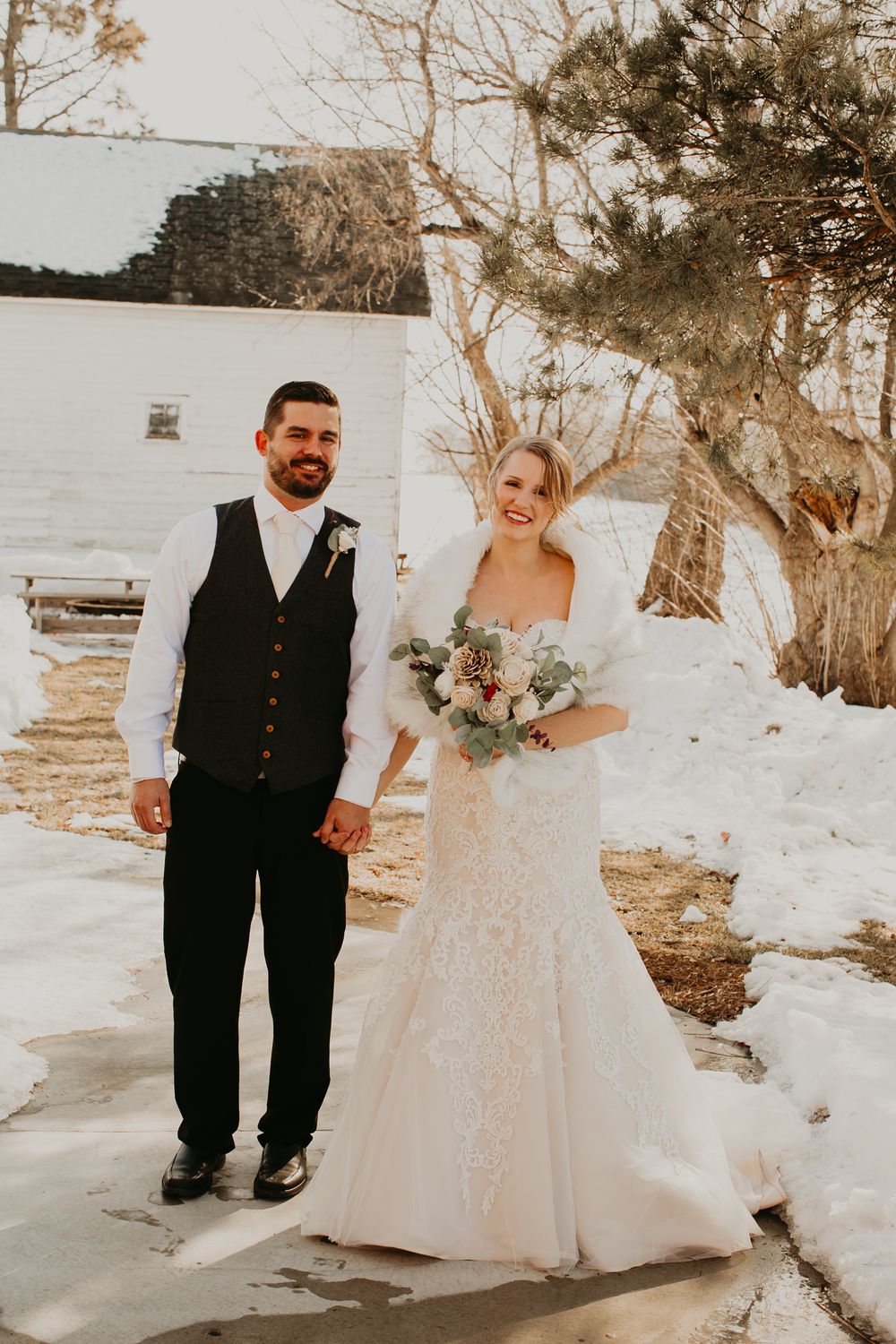 Bride and groom stand hand in hand outdoors in winter snow, bride in lace gown holding a floral bouquet.