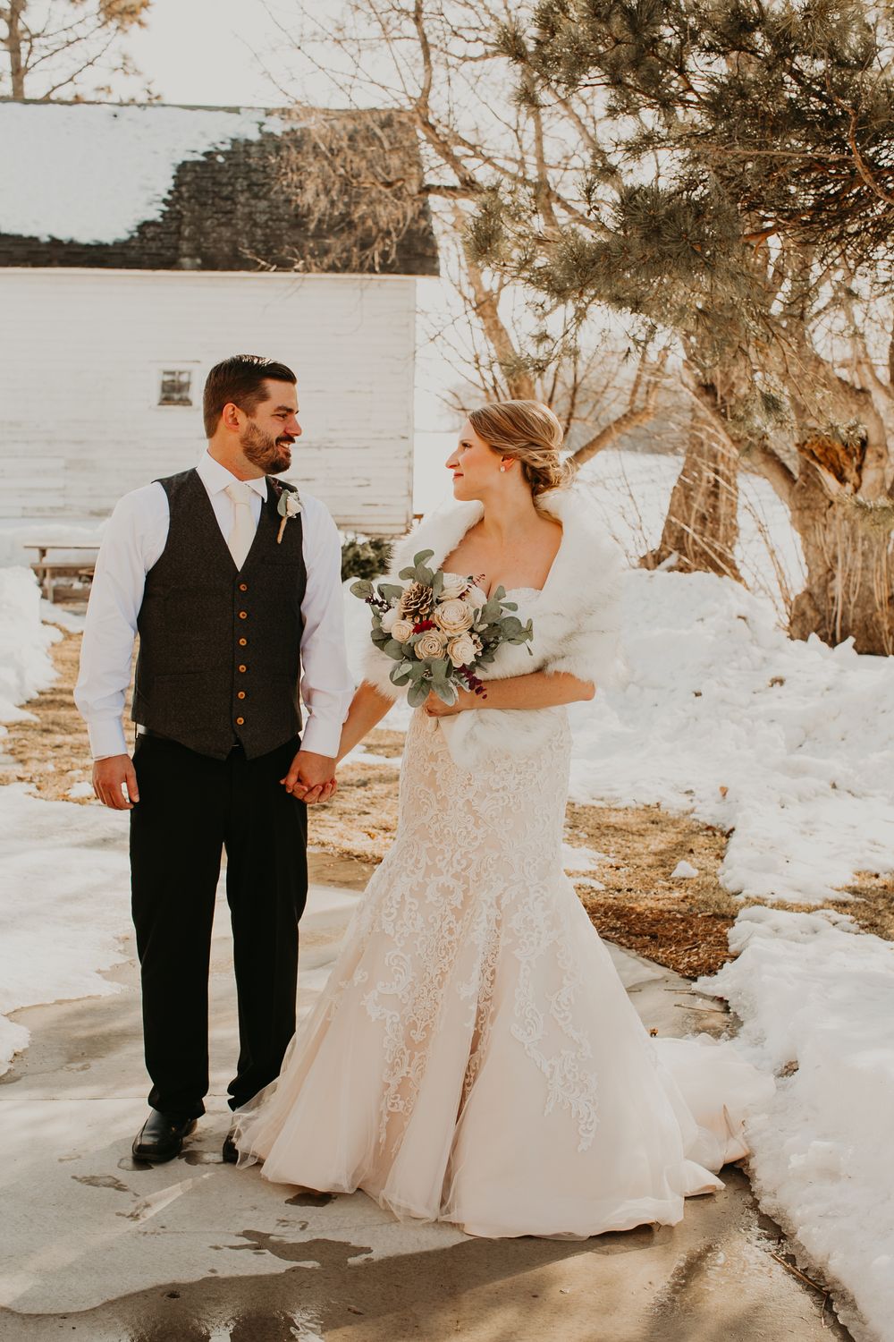 Bride and groom walk hand in hand through a snowy winter landscape, bride smiling in lace gown and fur wrap.