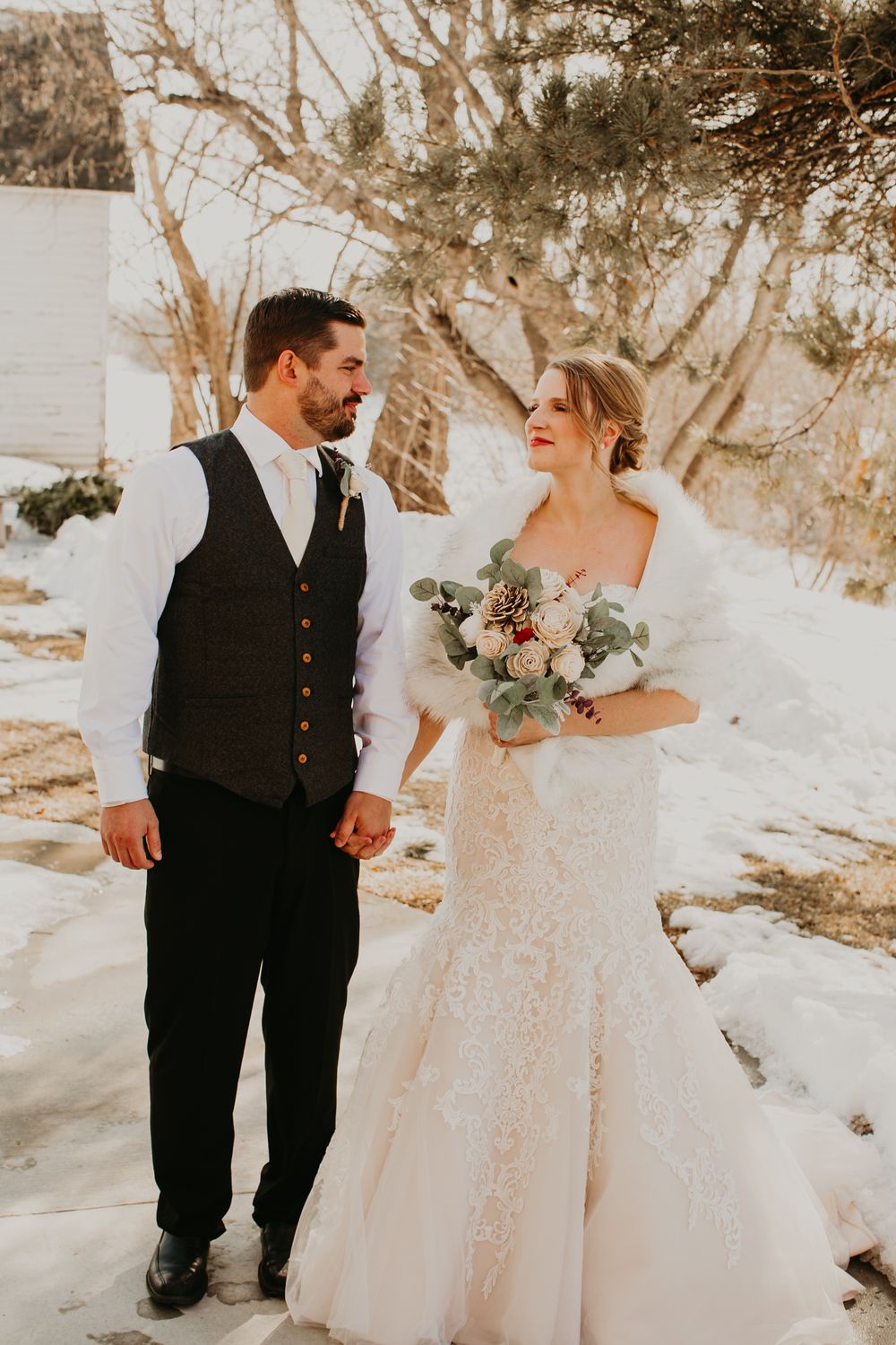 Bride and groom holding hands in snowy winter wedding setting, bride in lace gown with fur wrap holding bouquet.
