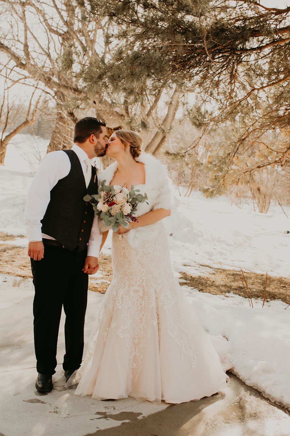 Winter wedding couple sharing a kiss outdoors in snow, bride in lace mermaid gown holding floral bouquet.