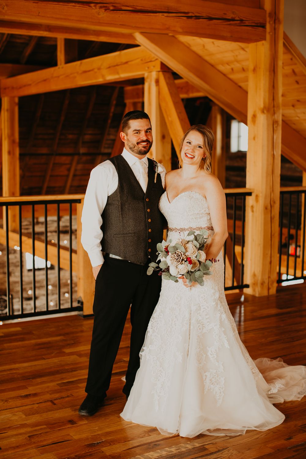 Smiling bride and groom posing inside rustic wooden barn venue, bride holding elegant floral bouquet.