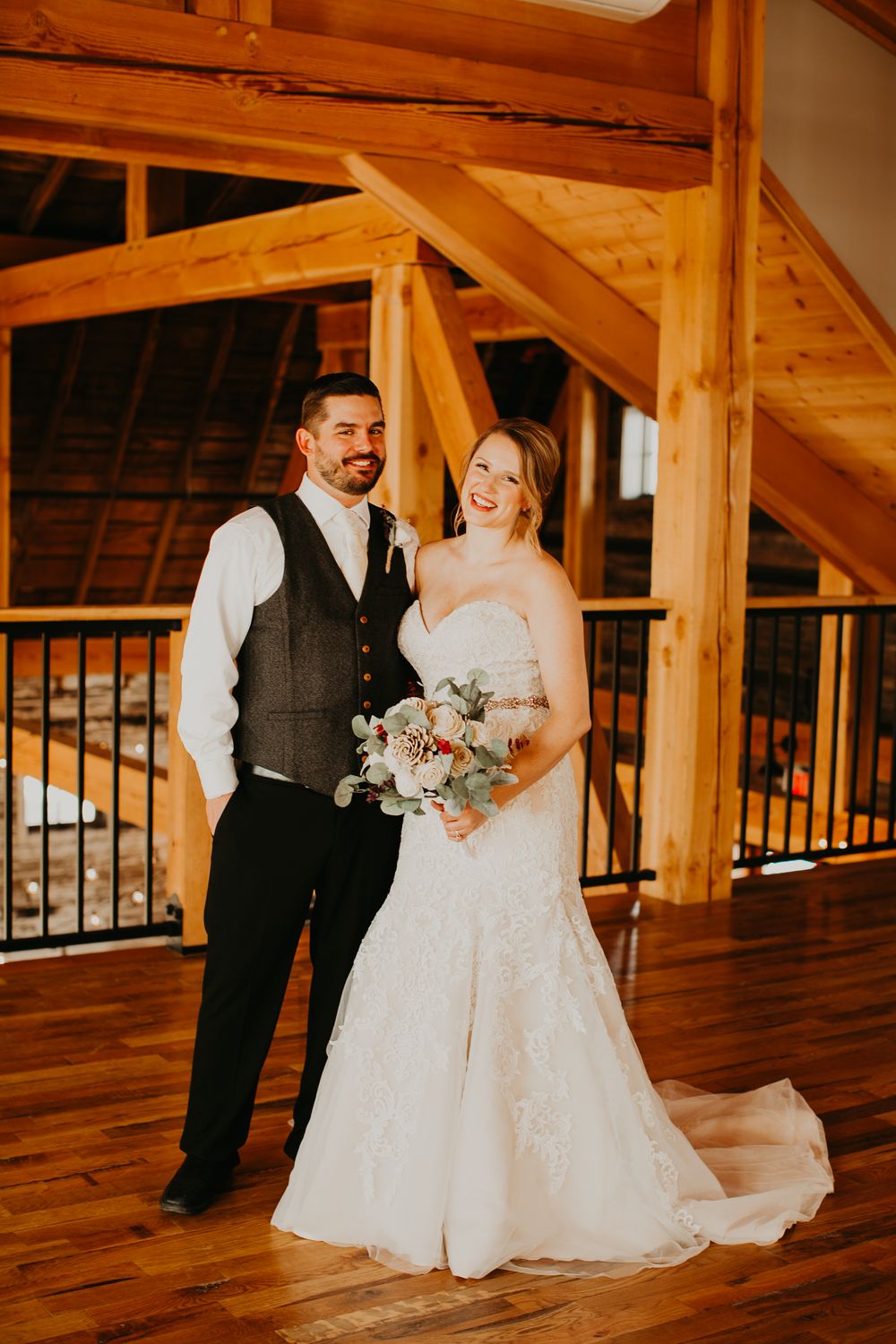 Happy wedding couple standing together in warm rustic barn, groom in grey vest beside bride in lace gown.