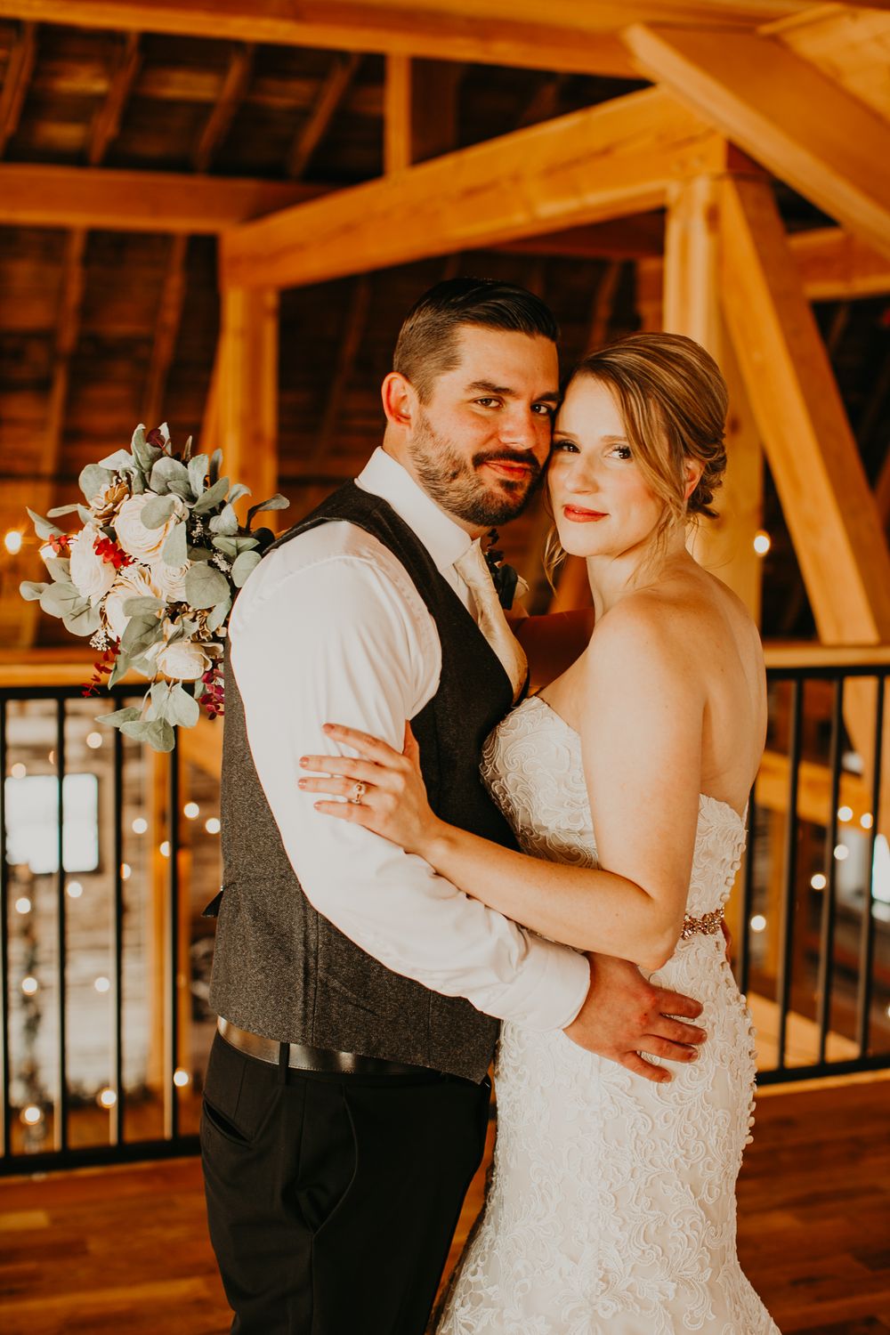 Groom embracing bride from behind in rustic barn venue with twinkling fairy lights in background.