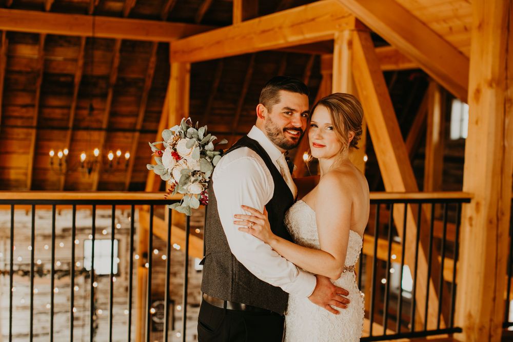 Wedding couple embracing in warmly lit rustic barn with chandelier and timber frame architecture.