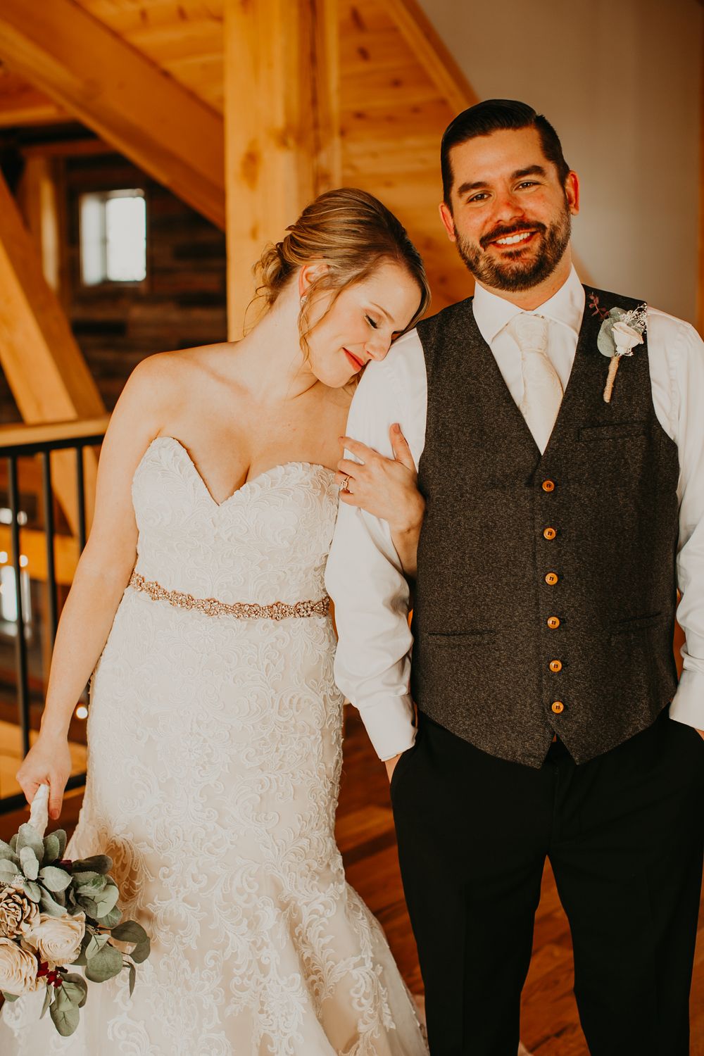 Smiling groom in gray vest with boutonniere stands beside bride in strapless lace wedding gown.