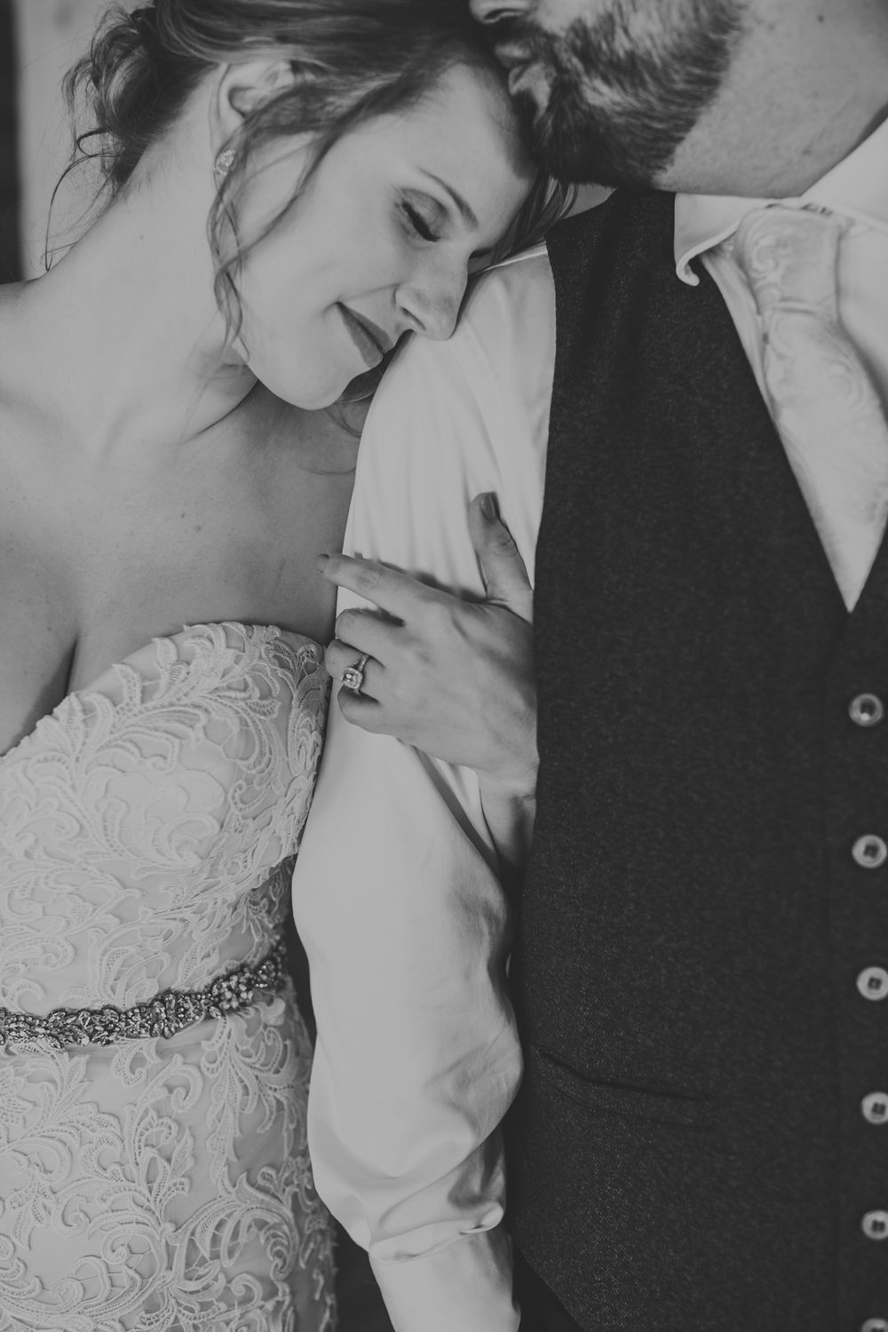 Black and white photo of bride in lace gown leaning on groom in vest, showcasing wedding ring.