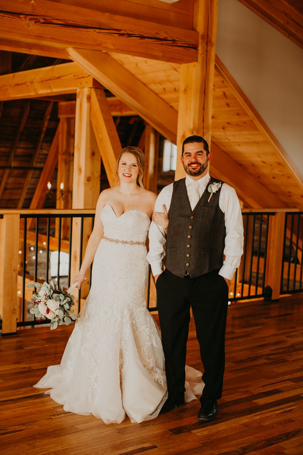 Bride and groom pose together in rustic timber frame barn, bride holding elegant floral bouquet.