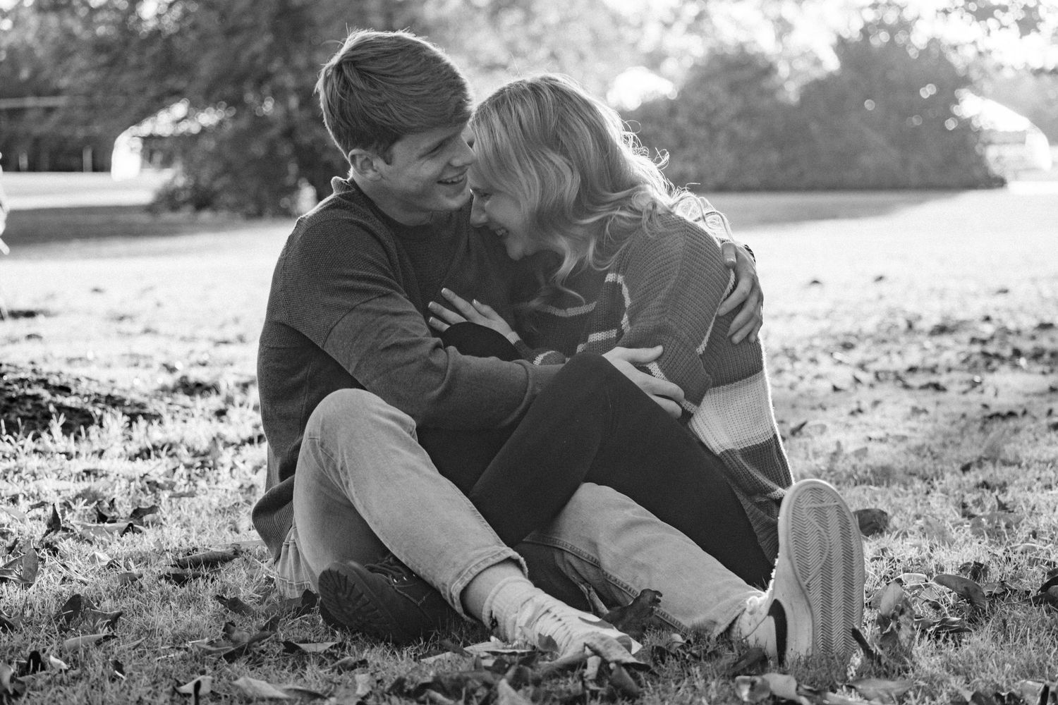 Young woman laughs as she leans into the embrace of her fiance while witting in a field