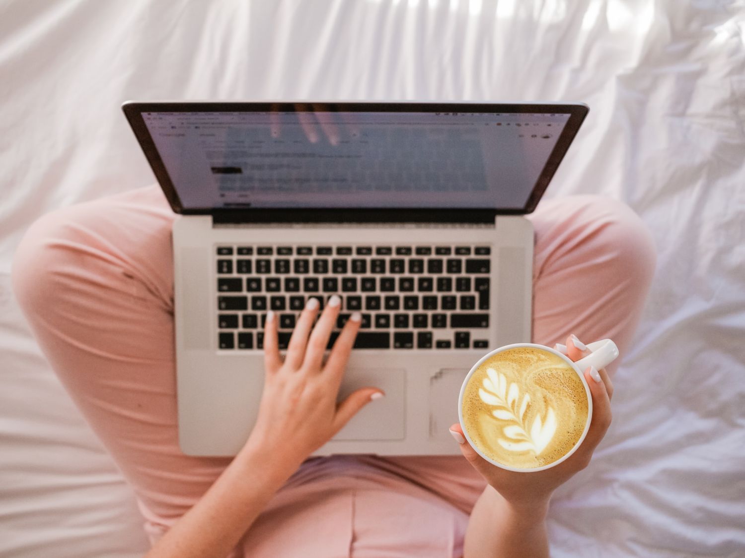 a woman in pink pants holding a laptop and latte on her bed