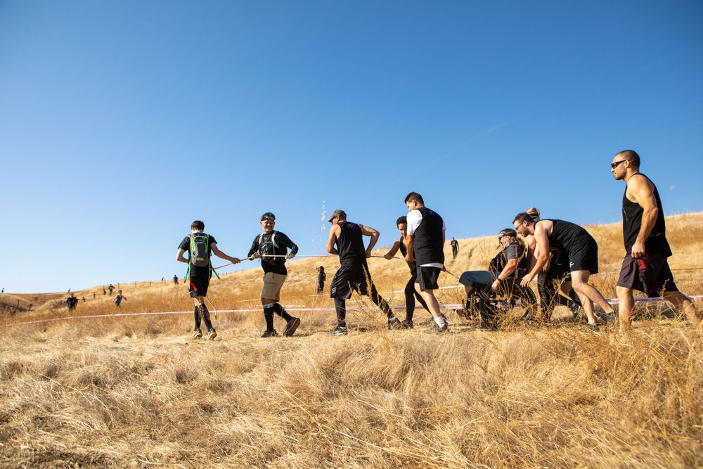 Group of participants running along dry hillside trail during outdoor obstacle course event.
