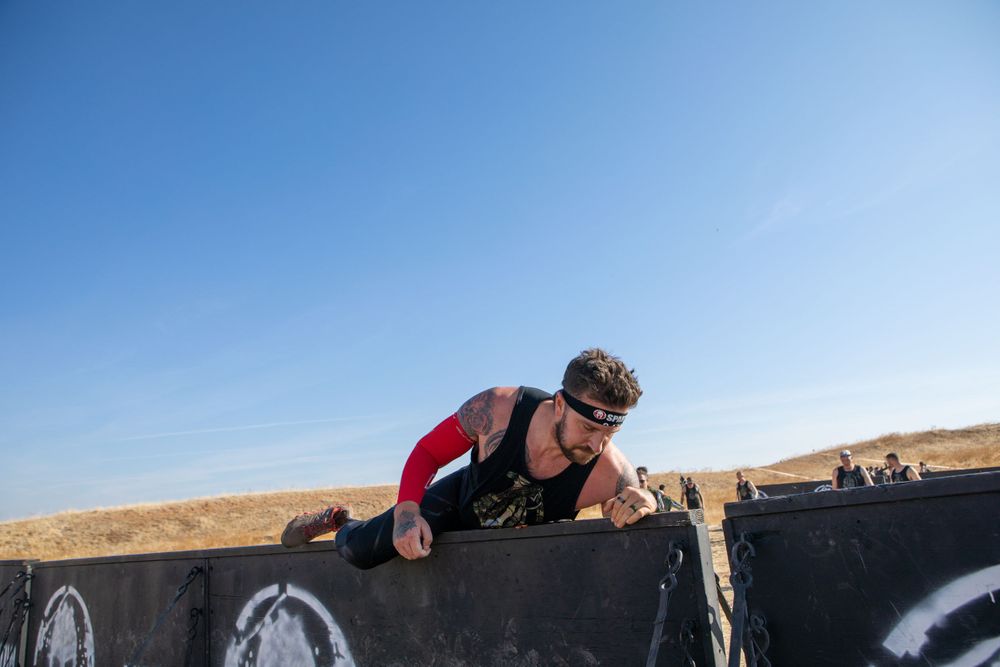 A participant climbs over a black wall obstacle during an outdoor competition.