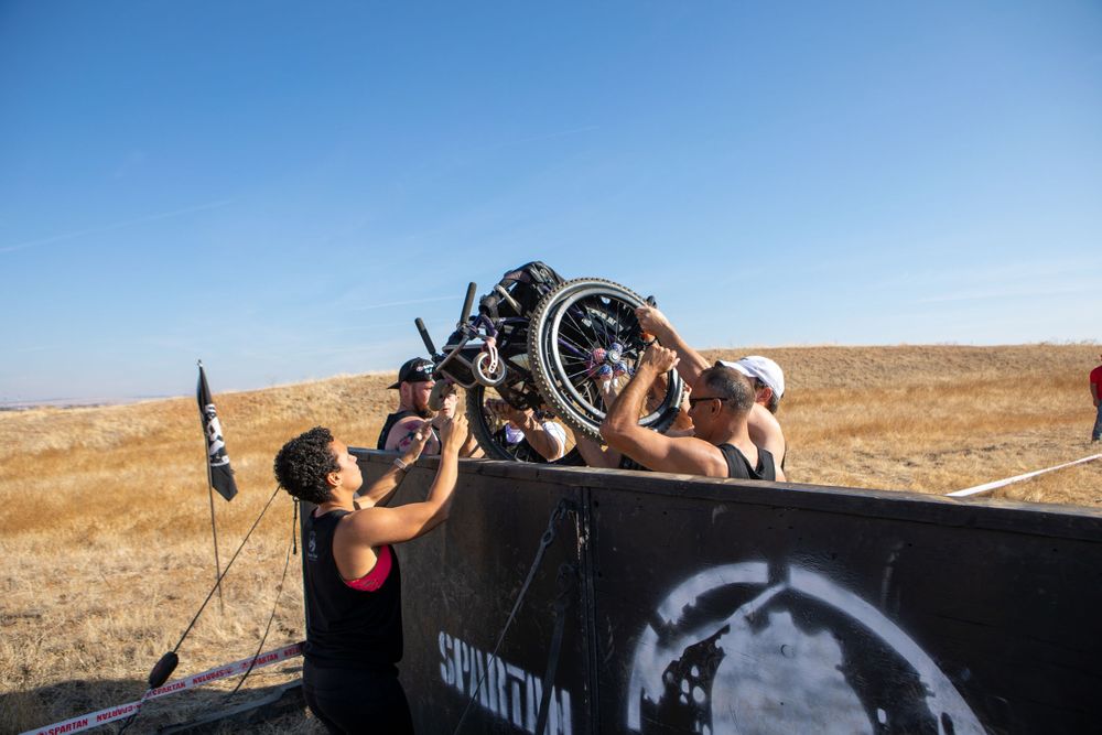 Team members lifting equipment over black obstacle barrier at outdoor event.