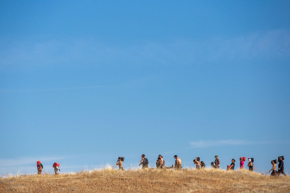 A line of people trek across a dry hilltop ridge against a bright blue sky.