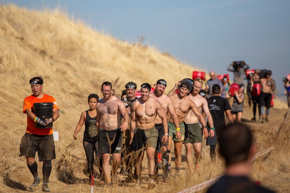 A group of participants run along a dusty hillside trail during an outdoor obstacle event.