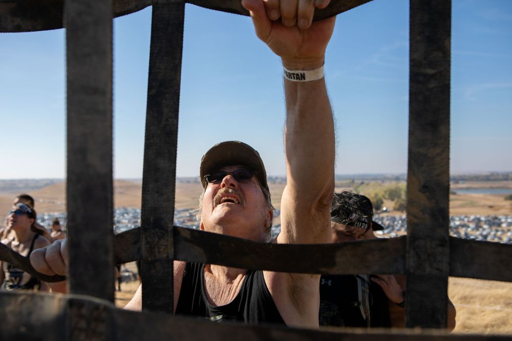 Competitor reaching through bars during an obstacle course challenge under bright sunlight.