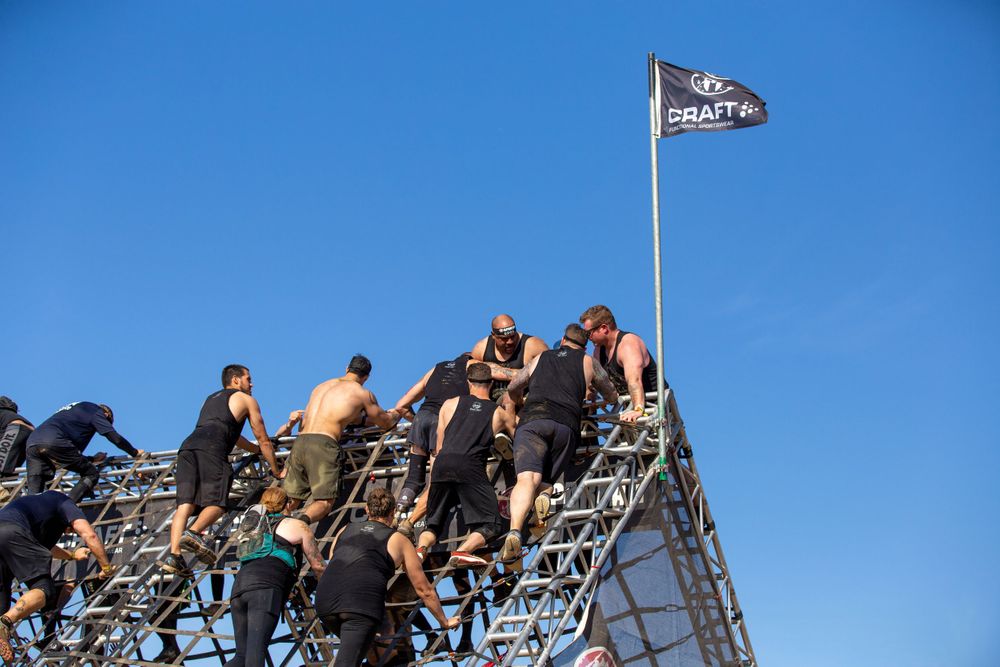 Participants scaling a metal framework structure with a flag at the top during an outdoor event.