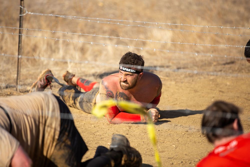 Participant crawling under yellow ropes at a muddy outdoor obstacle course.