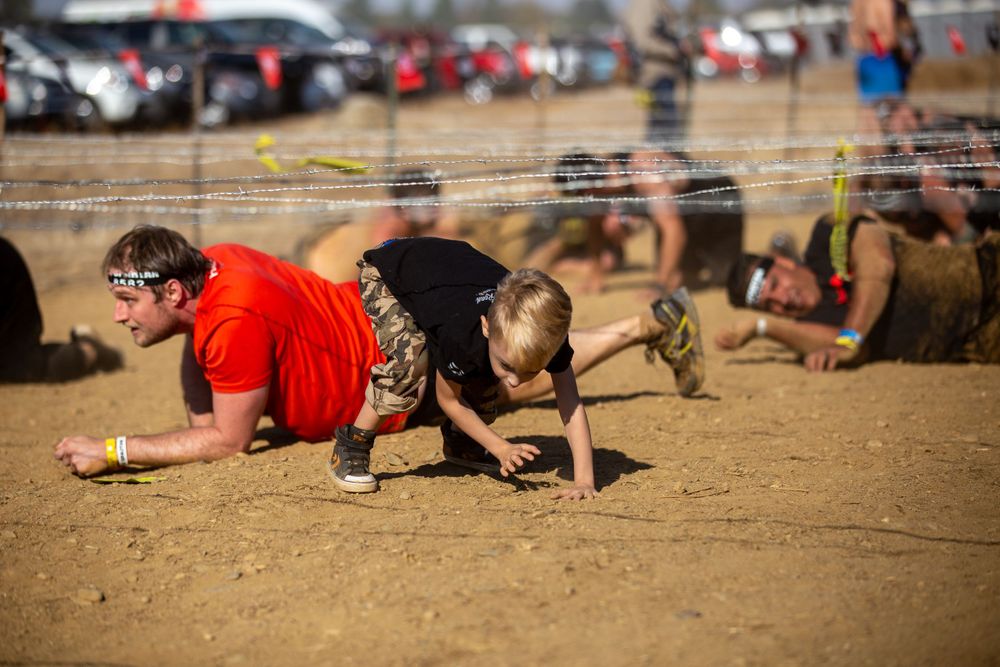Competitors crawling under low barriers during an obstacle course race in a dusty field.