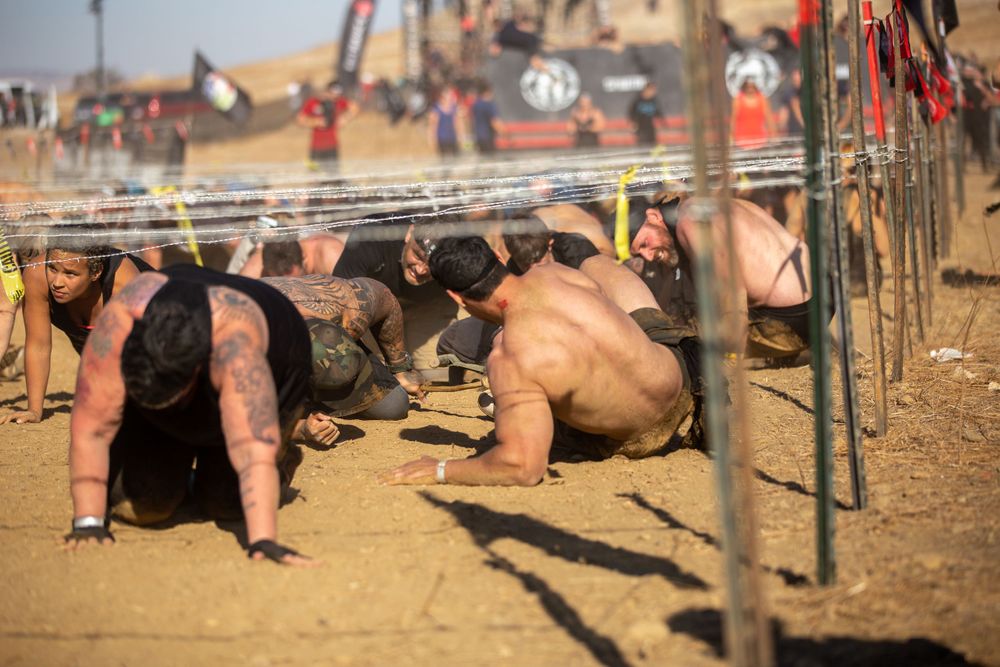 Athletes low crawling under wire obstacles at a dusty outdoor race course.