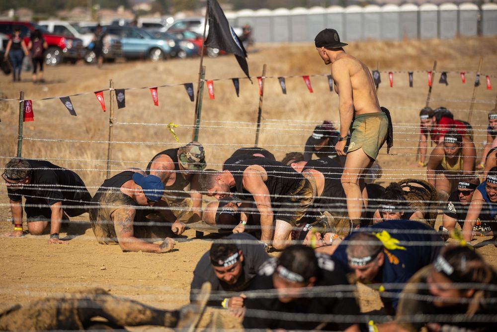Athletes crawling under obstacles at an outdoor endurance race with flags in the background.