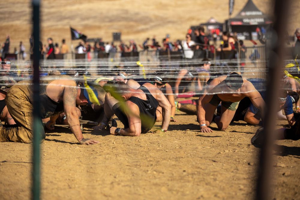 Athletes crawl under barbed wire obstacles during a challenging Spartan Race competition on dusty terrain.
