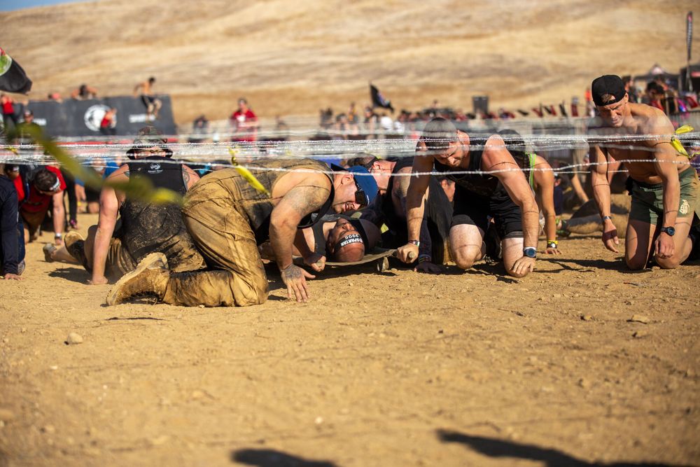 Competitors navigate under low barriers during a grueling obstacle course race in arid conditions.