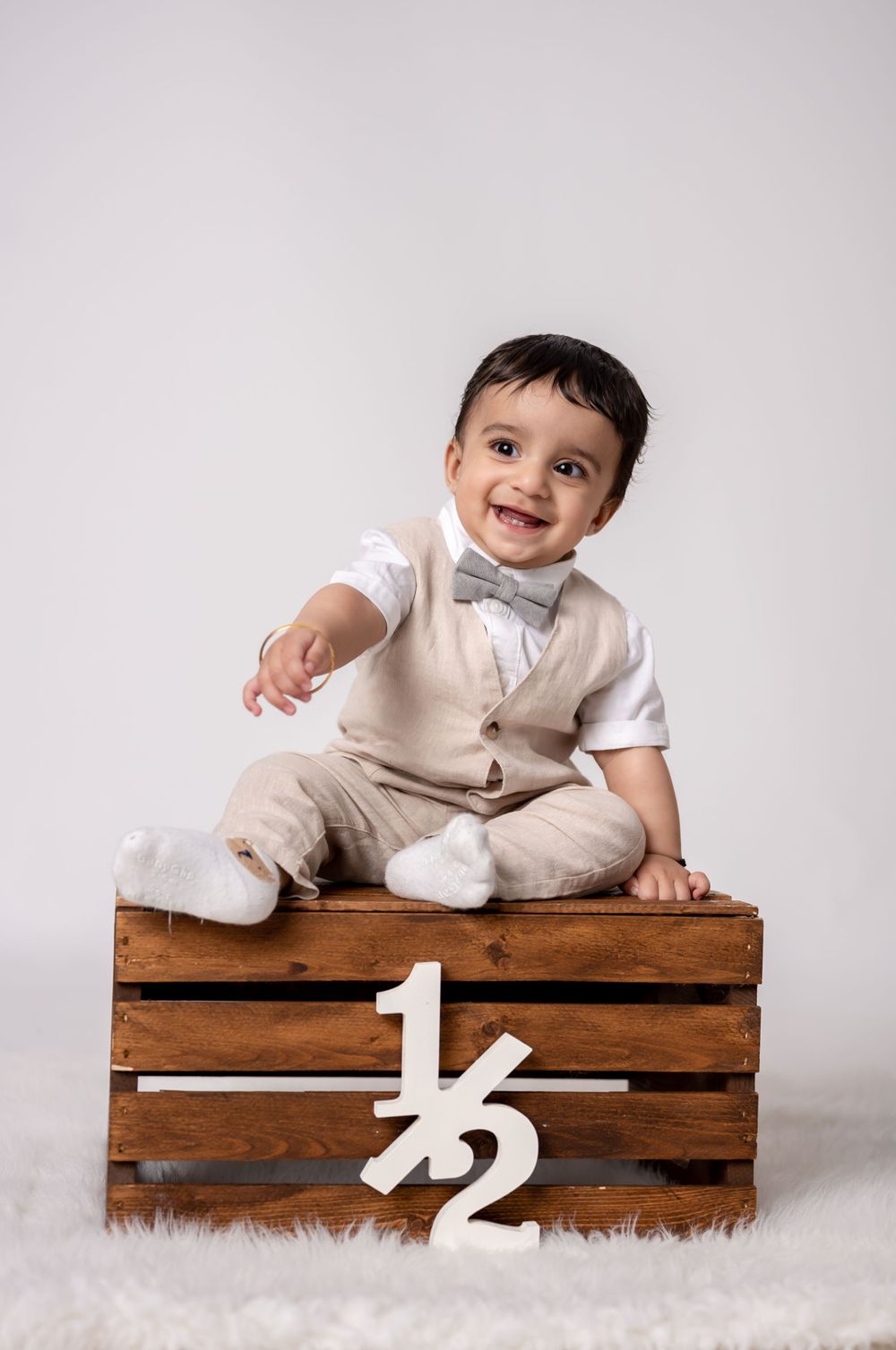 Baby in white formal outfit sitting on wooden crate with milestone numbers.