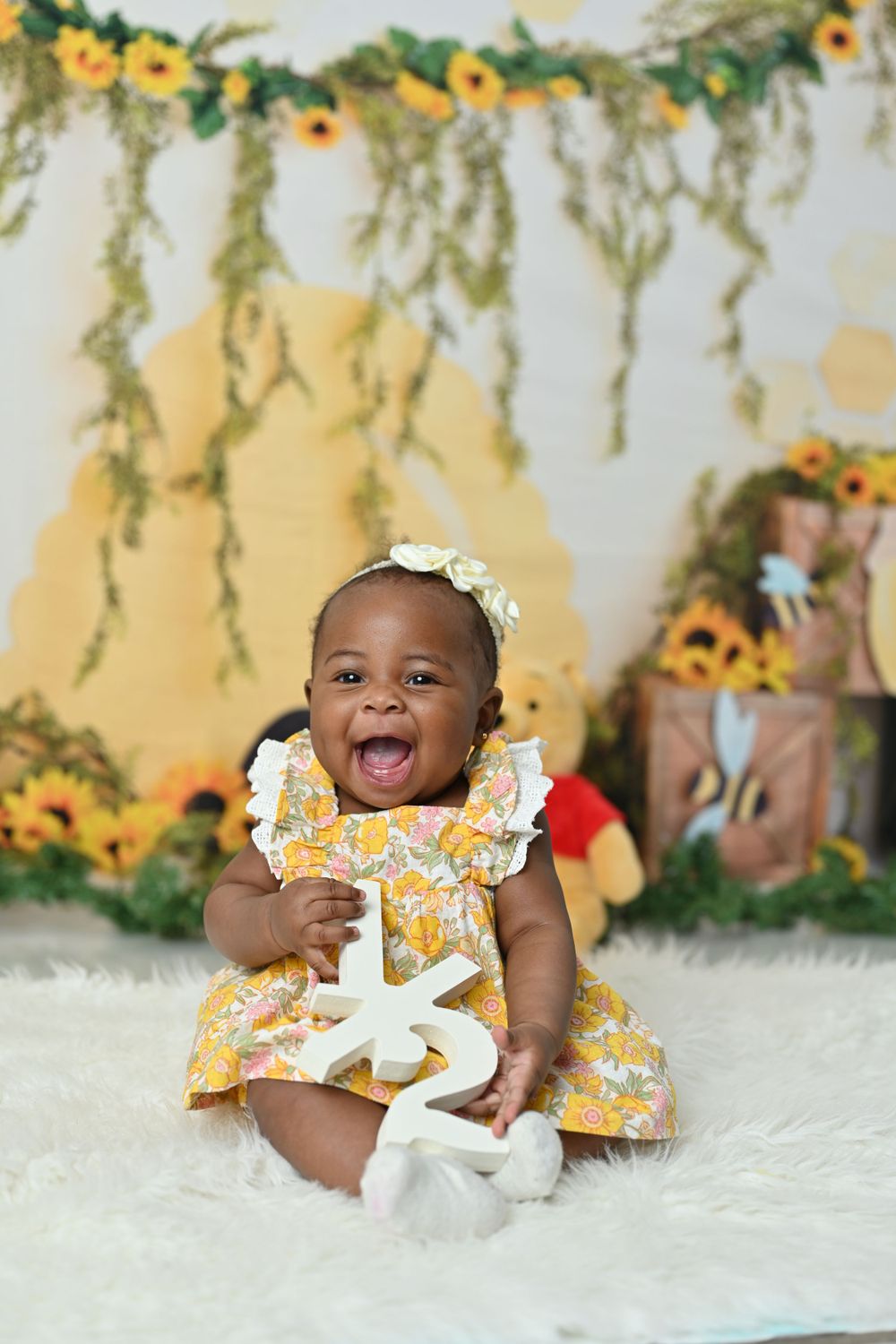 Baby in yellow floral dress lying on white rug with sunflower backdrop decorations.