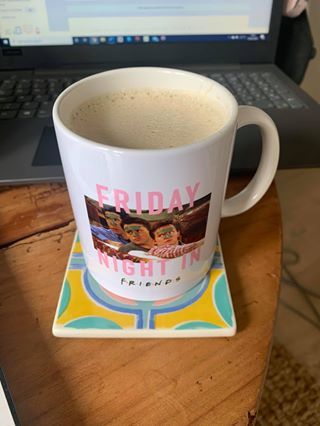 Coffee mug with Friends TV show theme sitting on colorful coaster near laptop keyboard.