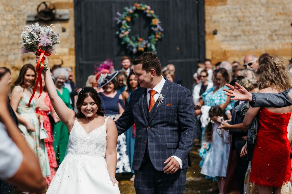 Wedding guests celebrate outdoors with colorful attire in front of a rustic brick wall with decorative wreath.