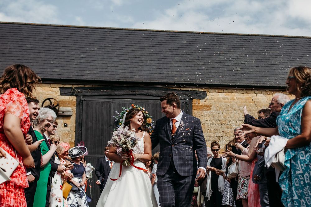 Outdoor wedding celebration with guests in colorful dresses throwing confetti near stone building.