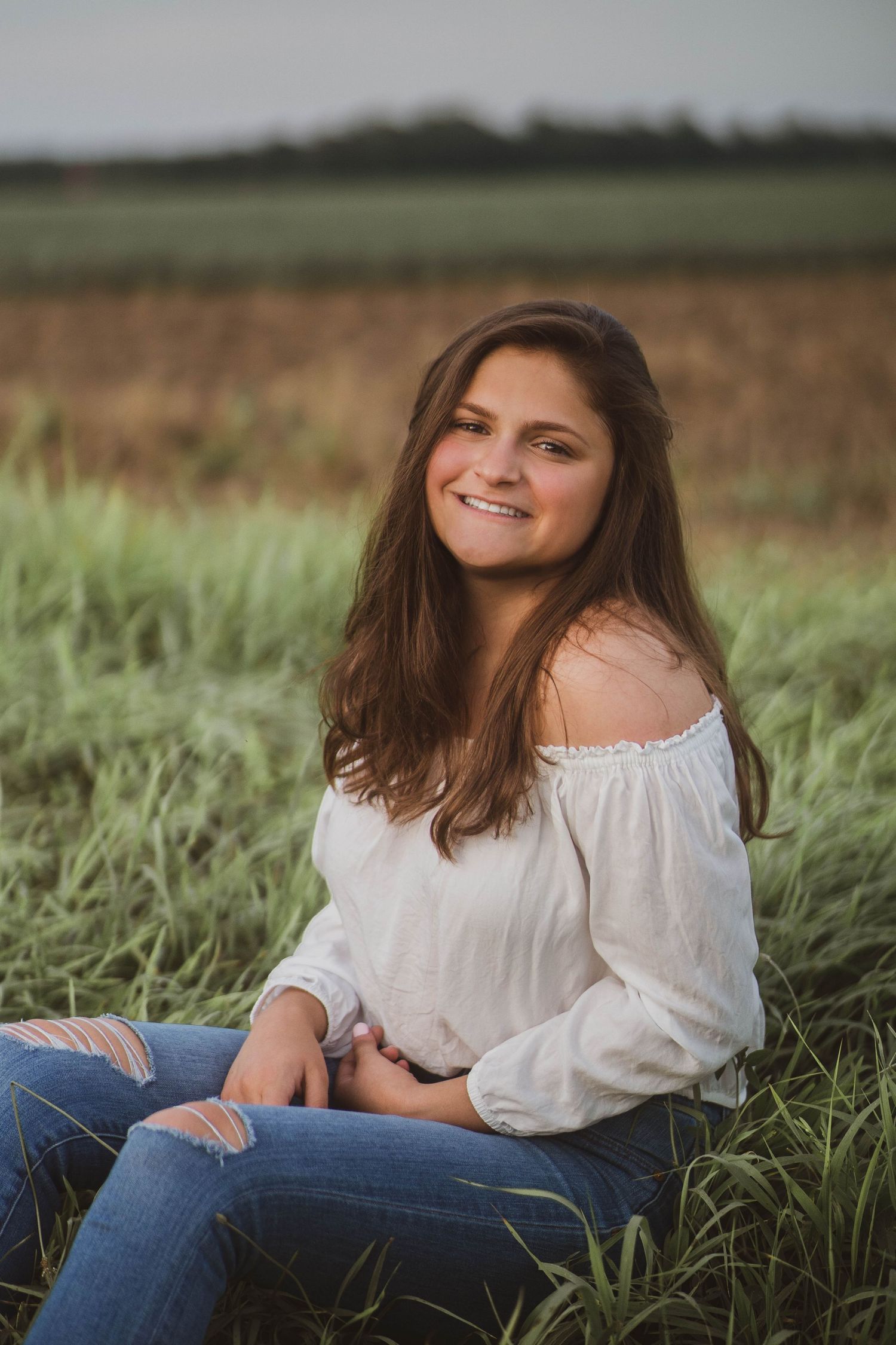Portrait of a person wearing a white off-shoulder top and jeans sitting in tall grass at sunset.