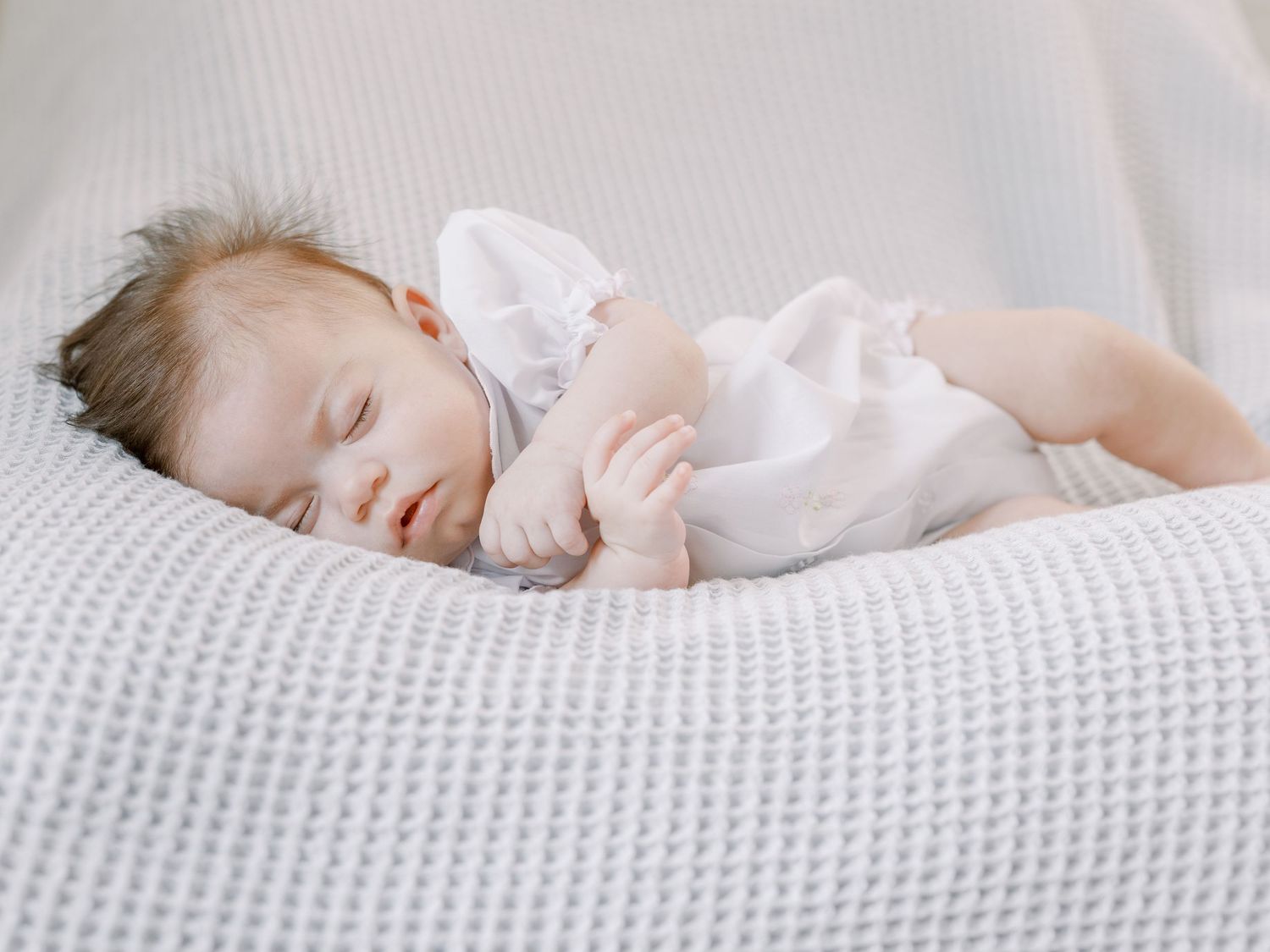 Peaceful sleeping newborn in white dress lying on textured white blanket.