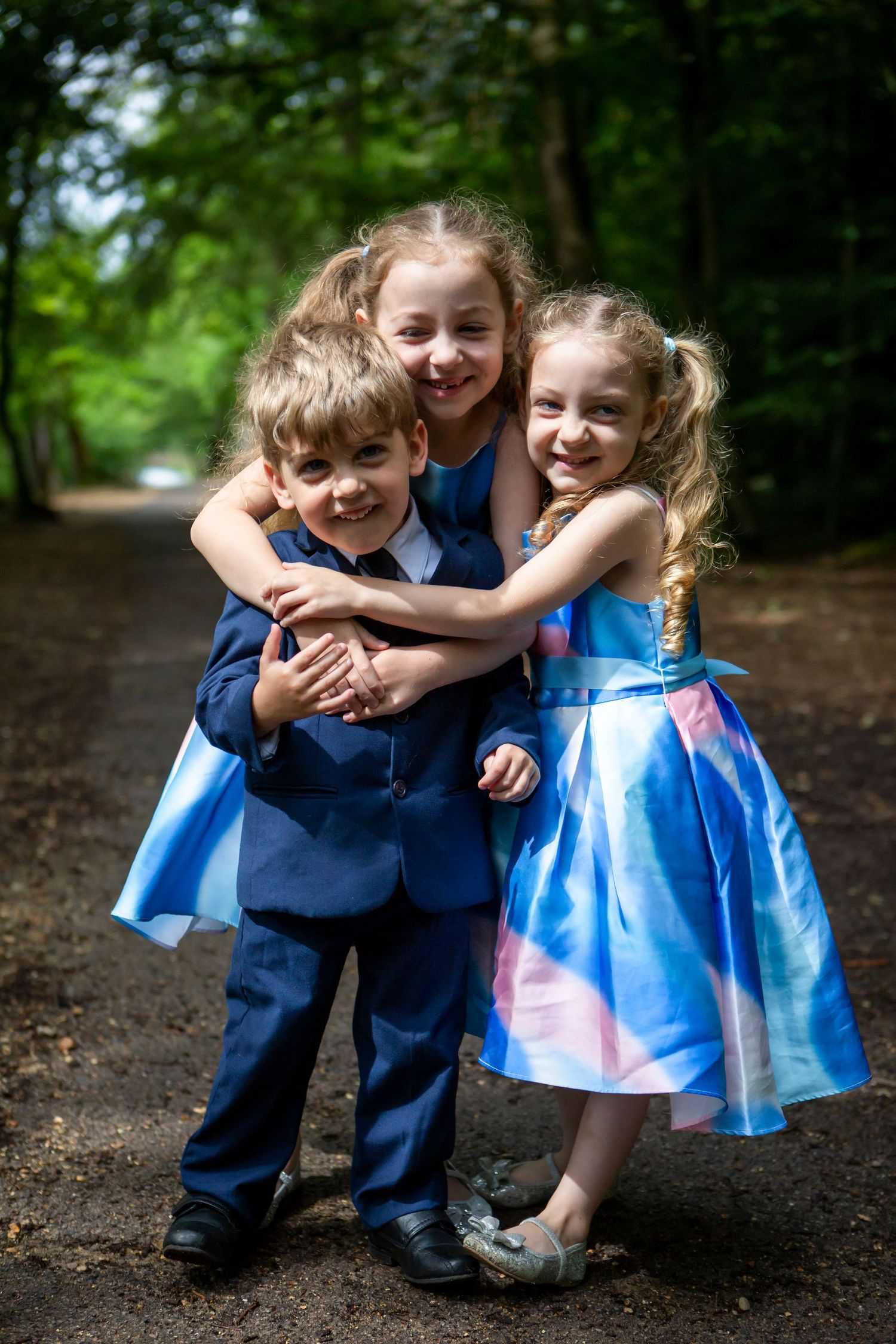 Three children in formal attire hugging on a woodland path.
